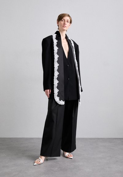 Zwarte oversized blazer met witte kanten rand, diepe V-hals, enkele knoop aan de voorkant, gecombineerd met wijde zwarte broek en witte sandalen met bandjes.