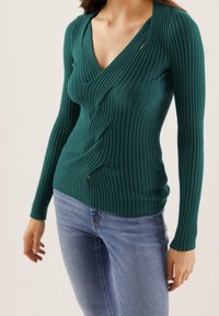 Femme portant un pull en maille côtelée vert foncé ajusté avec un design à l'avant torsadé et un jean bleu clair.