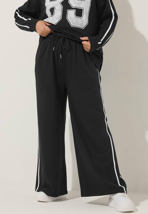 Pantaloni neri a gamba larga fatti di tessuto morbido, con strisce bianche ai lati, una vita elastica e una vestibilità rilassata. Abbinati a sneakers bianche.