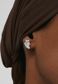Boucles d'oreilles en argent avec un design texturé mettant en valeur une pierre précieuse claire, portées sur l'oreille, sur fond de tissu marron.