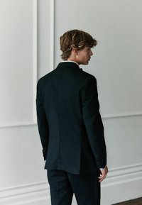 Schwarzer Anzug mit schmaler Passform, ausgestattet mit einem einzigen Schlitz und geknöpften Manschetten. Glatter Stoff mit dezentem Glanz, getragen mit einem frischen weißen Hemd.