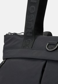 Sac en tissu noir avec une épaisse sangle cousue portant le texte "HUGO", fermeture à glissière et un anneau en D noir cousu sur fond clair.