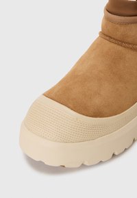 Tan suède enkellaars met een afgeronde beige rubberen zool en een gestructureerde teenkap. Geëlasticeerde kraag en gestikte accenten benadrukken het ontwerp.