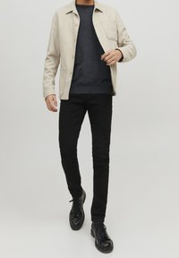 Beige Wollmischung Knopfhemdjacke mit einer Brusttasche, über einem dunklen Pullover getragen und kombiniert mit schwarzen Slim-Fit-Jeans und schwarzen Schnürstiefeln.
