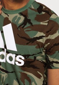Groene camouflage t-shirt met een ronde halslijn. Kenmerkt een groot wit 'adidas' logo op de voorkant. Zachte stof met afwisselende groene en bruine patronen.