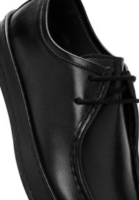 Zapato de cuero negro con puntera redonda, textura suave y costuras en contraste. Presenta dos ojetes para los cordones y una suela de perfil bajo.