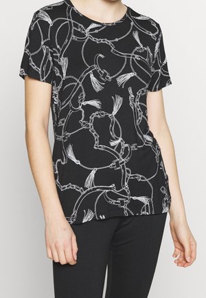 Personne portant une chemise noire à manches courtes avec un motif de chaînes blanches, de glands et de ceinture, associée à un pantalon noir.