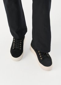 Schwarze Wildledersneaker mit goldenen Ösen und schwarzen Schnürsenkeln, ausgestattet mit einer runden Zehenpartie und einer weißen strukturierten Sohle. Getragen mit dunklen Hosen.
