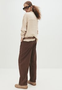 Frau mit lockigem Haar in oversized beiger Bluse, braunen weiten Hosen, hellbraunen Sneakers, trägt große dunkle Sonnenbrillen, steht vor weißem Hintergrund.