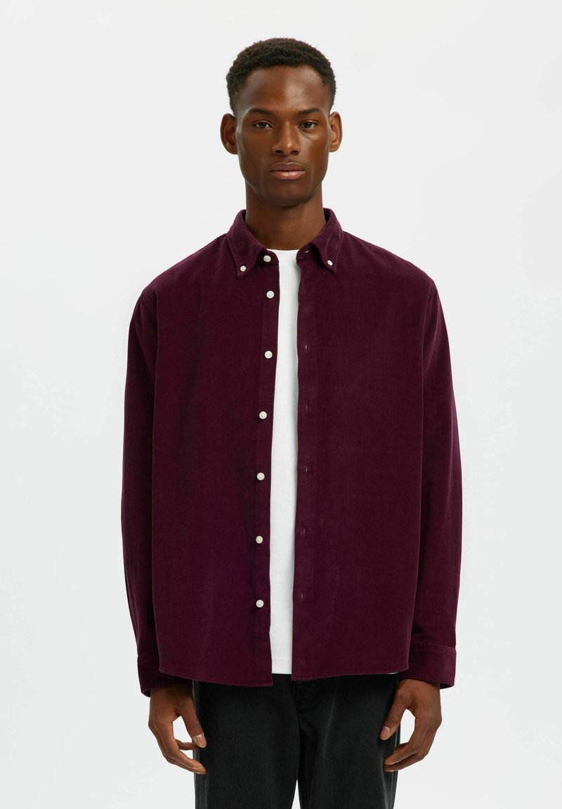 Bordeaux corduroy blouse met een button-down kraag, witte knopen, lange mouwen, zakken, gedragen over een wit T-shirt, in combinatie met donkere jeans.