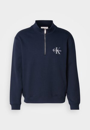 Tummansininen, vetoketjullinen huppari, joka on valmistettu pehmeästä kankaasta. Siinä on korkea kaulus, ribbaatut hihansuut ja rintahilineen logo vastakkaisissa väreissä.