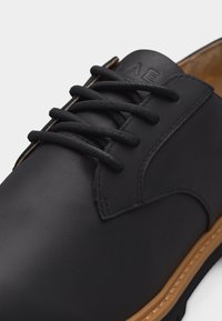 Μαύρο οξφορδ παπούτσι από δέρμα με στρογγυλά μαύρα κορδόνια, ορατή ραφή και λεπτομέρεια σε σόλα χρώματος ταμπά σε λευκό φόντο.