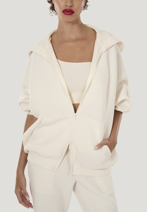 Vrouw draagt een off-white hoodie met ritssluiting over een bijpassende crop top en broek, met een hand in de zak, neutrale achtergrond.