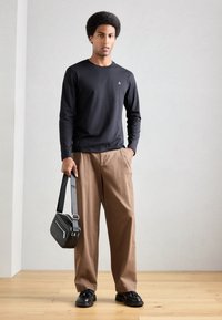 Camisetă neagră cu mâneci lungi, pantaloni maro lejeri, geantă de umăr texturată neagră și pantofi negri, stând pe un podea din lemn, în fața unui perete deschis.