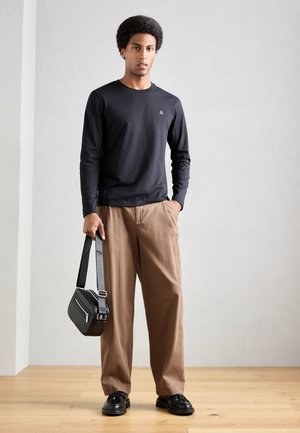 Zwart langemouw shirt, bruine losse broek, zwarte gestructureerde schoudertas en zwarte schoenen, staand op een houten vloer tegen een lichte muur.