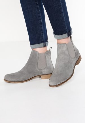 Personne portant un jean bleu foncé retroussé et des bottines en daim gris à talons en bois bas sur un sol blanc.