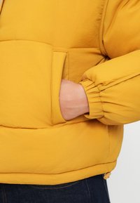 Kollane polsterdatud jakk pehme tekstuuriga, küljetasku, elastsete mansettide ja gathered-äärise detailiga.