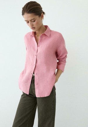 Vrouw met een losvallend roze overhemd met opgerolde mouwen en donkergroene broek, staand met handen in de zakken tegen een witte achtergrond.