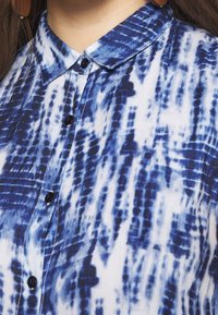 Blusa blu e bianca a fantasia con colletto, chiusura a bottoni e un tessuto leggero con un design testurizzato simile al tie-dye.