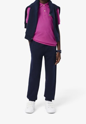 Pantalon de survêtement bleu marine, polo rose et un pull bleu marine posé sur les épaules. Baskets blanches avec fermetures par scratch. Tissu lisse, design décontracté.