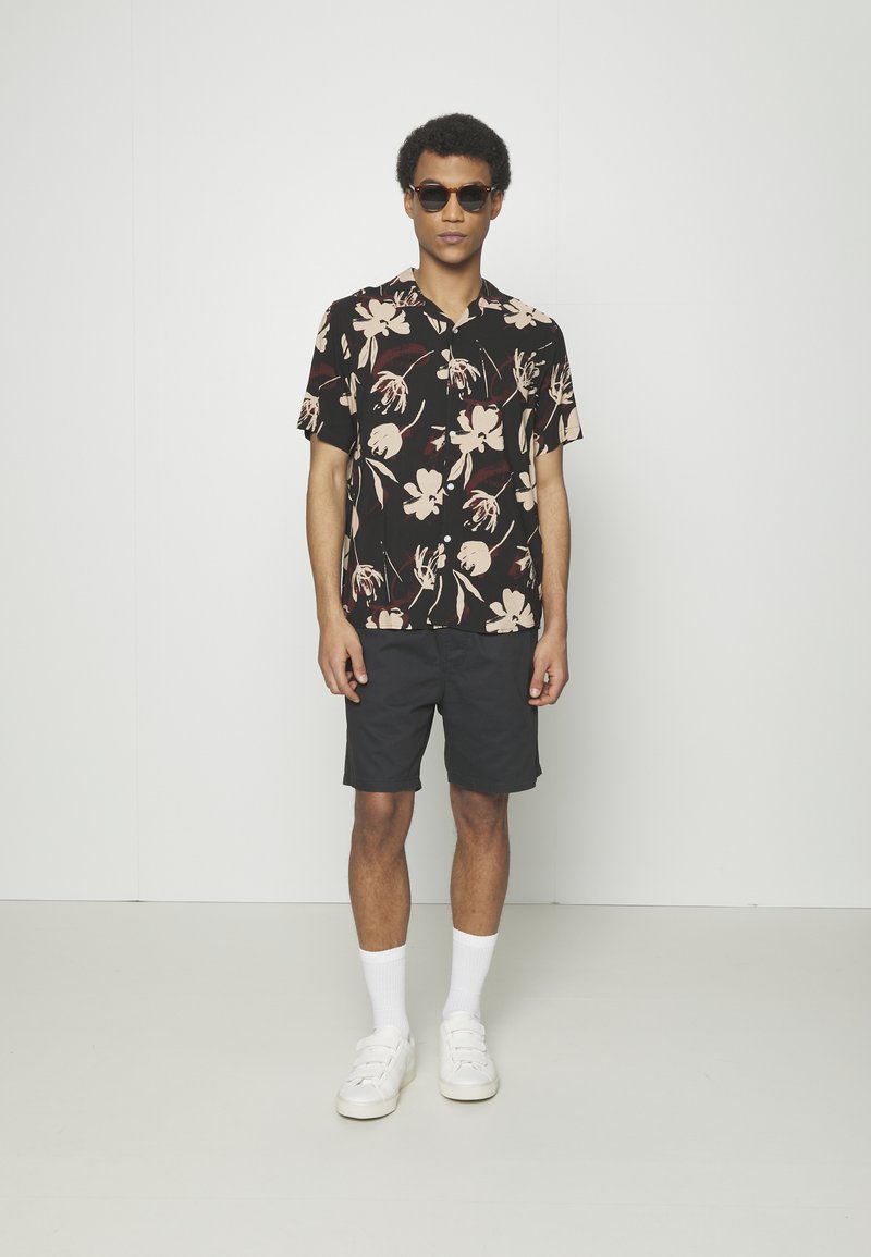 Chemise noire à manches courtes avec un motif floral en crème et bordeaux, associée à un short foncé et des baskets blanches, accessoirisée avec des lunettes de soleil.