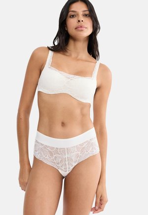 Kvinde iført hvid blonde-bh og matchende blondeundertøj, stående mod en lys, ensfarvet baggrund.