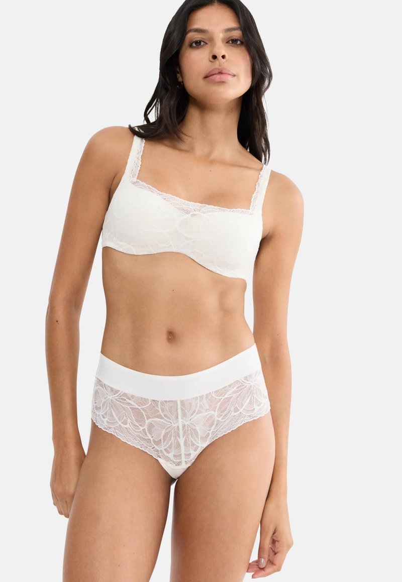 Γυναίκα που φοράει λευκό δαντελωτό σουτιέν και ταιριαστό δαντελωτό εσώρουχο, στέκεται μπροστά σε ένα απλό φωτεινό φόντο.