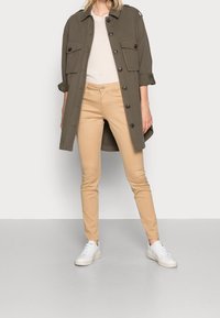 Lättviktig olivgrön knäppjacka med två framfickor, tillsammans med beige åtsittande byxor och vita sneakers på en neutral bakgrund.