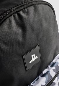 Sac à dos noir en tissu texturé, avec un patch logo PlayStation bien en vue et une sangle à motif camouflage.