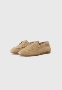 Beige Wildleder-Slipper mit niedrigem Profil, runder Zehenpartie und dekorativer Naht. Mit Schnürung und Metallösen an den Seiten.