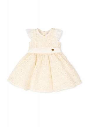Vestito per bambina senza maniche color crema con pizzo floreale traforato, spalle arricciate e una cintura bianca con un piccolo ciondolo a forma di cuore dorato.