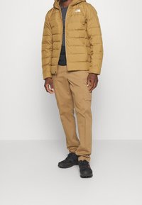 Brun puffjacka med horisontell quiltning, svart t-shirt, beige cargo-byxor och svarta sneakers. Anmärkningsvärda detaljer som huva och dragkedja.