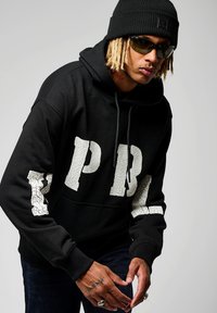 Sweat à capuche noir avec de grandes lettres blanches fissurées "PBL" sur les manches. Le matériau semble doux. Porté avec un bonnet noir et des lunettes de soleil.