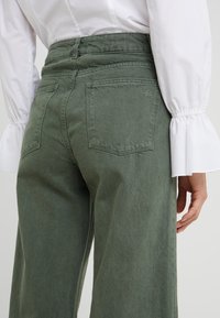 Henkilö, jolla on ylhäältä vyötä puristavat oliivinvihreät leveälahkeiset housut takataskuilla ja valkoinen pitkähihainen paita, jossa on levenevät hihansuut.