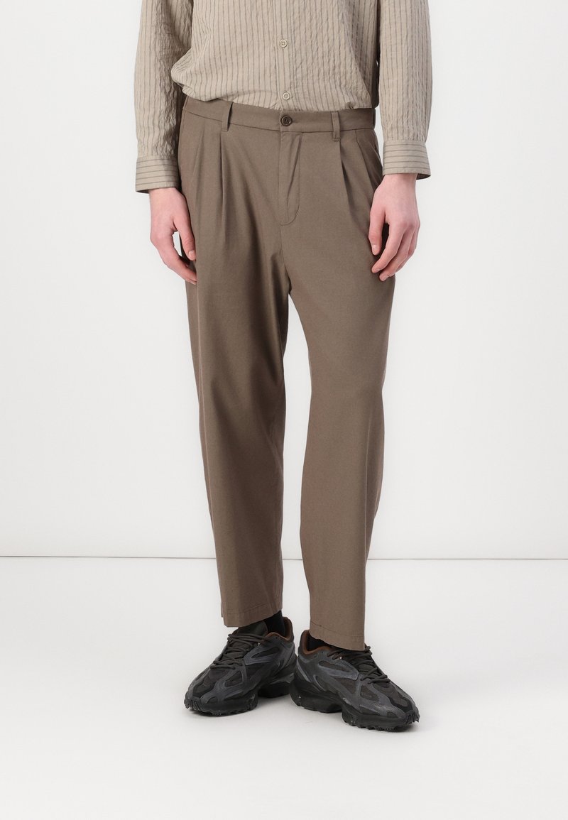 Personne portant une chemise beige à rayures, un pantalon marron plissé et des baskets noires, debout sur un fond blanc uni.