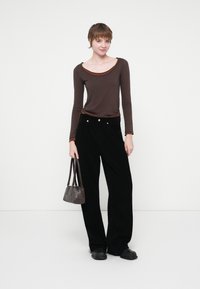 Camicia marrone a maniche lunghe con scollo a barca, abbinata a pantaloni ampia in corduroy neri. Porta una piccola borsa scura. Calzature nere.