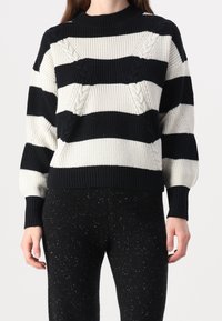 Suéter de punto con rayas horizontales en blanco y negro, con patrones de trenzado texturizado. Cuello y puños de canalé. Combinado con pantalones negros con manchas.