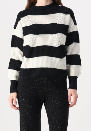 Gestrickter Pullover mit schwarz-weißen horizontalen Streifen, der strukturierte Kabelmuster aufweist. Ribbelkragen und -manschetten. Kombiniert mit schwarz-gesprenkelter Hose.