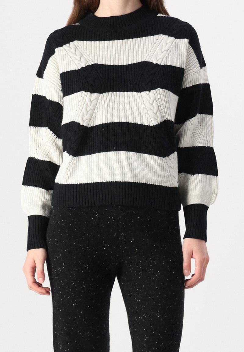 Suéter de punto con rayas horizontales en blanco y negro, con patrones de trenzado texturizado. Cuello y puños de canalé. Combinado con pantalones negros con manchas.