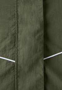 Gros plan sur un tissu vert olive avec des coutures verticales et un détail de bordure diagonale blanche.