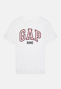 T-shirt alb din bumbac, cu inscripția "GAP" în roșu cu contur negru și "ROME" în negru îngroșat. Maneci scurte și design cu gât rotund.