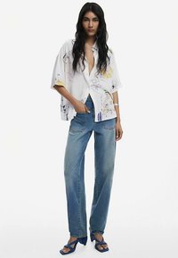 Camicia bianca oversize a maniche corte con stampe floreali colorate, abbinata a jeans in denim blu a vita alta e sandali blu con cinturini.