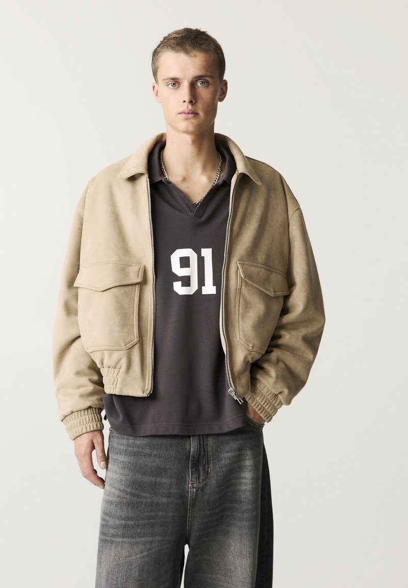 Beige Wildleder-Bomberjacke mit zwei Fronttaschen, kombiniert mit einem dunkelgrauen Shirt mit der weißen Nummer '91' und locker sitzenden grauen Jeans.