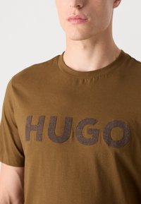 Barna pamut póló kerek nyakkal és rövid ujjakkal, amelyen egy nagy, textúrált "HUGO" logó található sötétebb barnában.