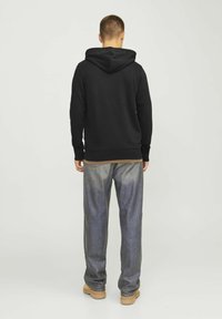 Black hoodie with a front pocket and drawstring hood, paired with loose-fitting grey jeans and tan boots, viewed from the back.