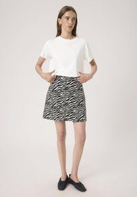 Junge Frau, die einen schwarz-weißen Zebramuster-Rock zurechtrückt, trägt ein schlichtes weißes T-Shirt und schwarze Ballerinas und steht vor einem hellen Hintergrund.