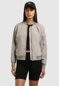 Jeune femme portant une veste bomber beige sur un crop top noir, un short de cyclisme noir, et une casquette noire avec un logo blanc.