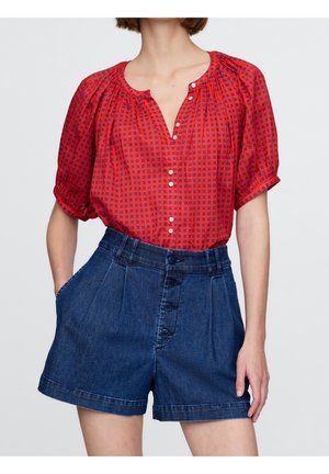 Kobieta ubrana w czerwoną, wzorzystą bluzkę z krótkimi bufkami oraz wysokowodne ciemne szorty jeansowe z przodu zapinane na guziki i posiadające kieszenie.