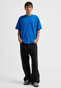 Blå överdimensionerad t-shirt i bomull med en tryckt logotyp på bröstet, matchad med svarta baggybyxor och grå sneakers.