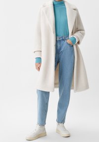Persona con un abrigo de teddy blanco sobre un suéter de cuello alto azul, jeans de un azul claro y zapatillas altas blancas, de pie con una mano en el bolsillo.